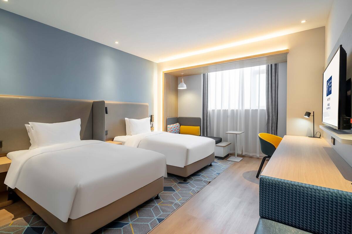 A hotel room with two beds, a desk, a chair, a TV, and a window with curtains.