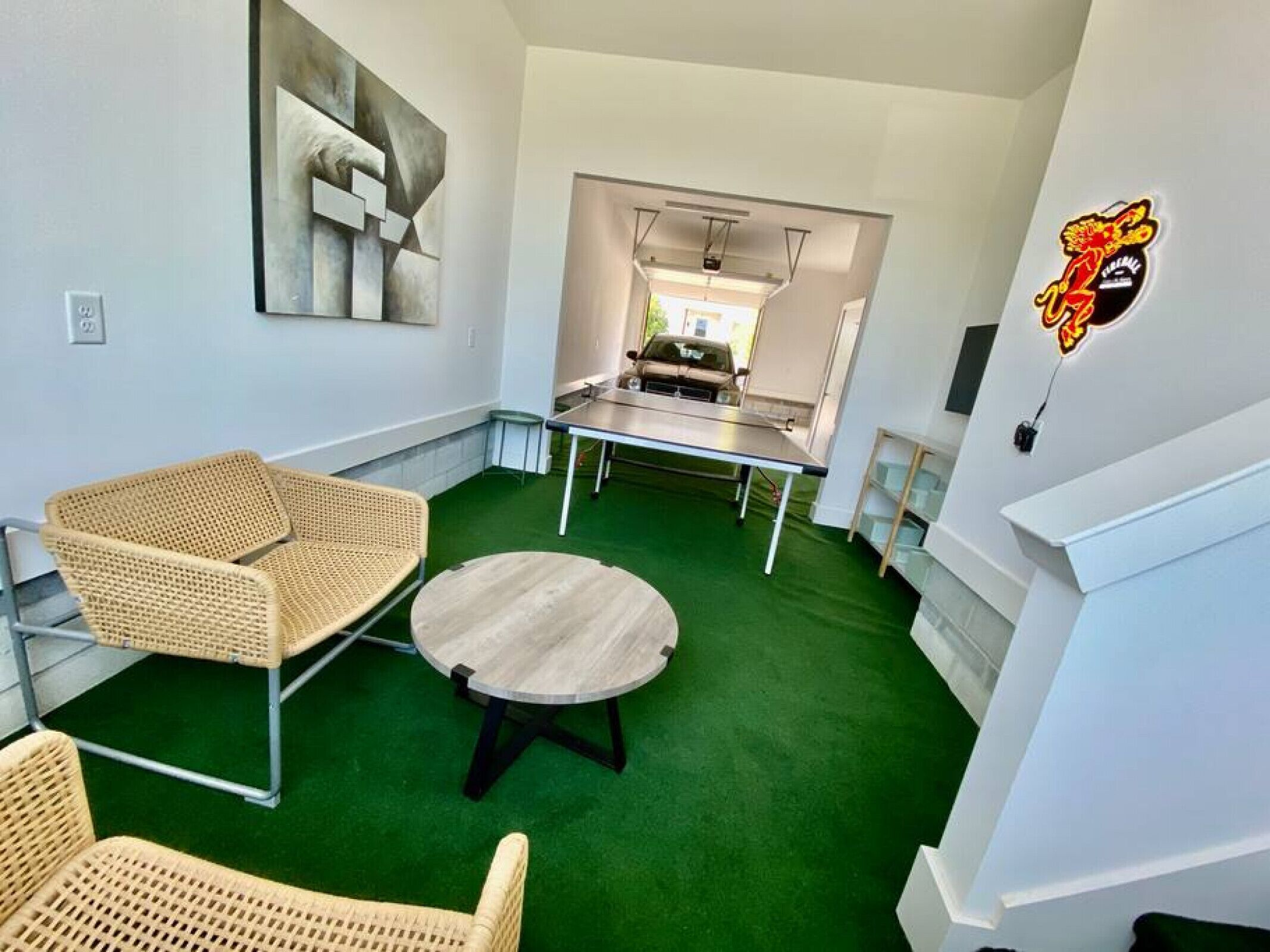 A room with a green carpet, wicker chairs, a round table, and a painting on the wall.
