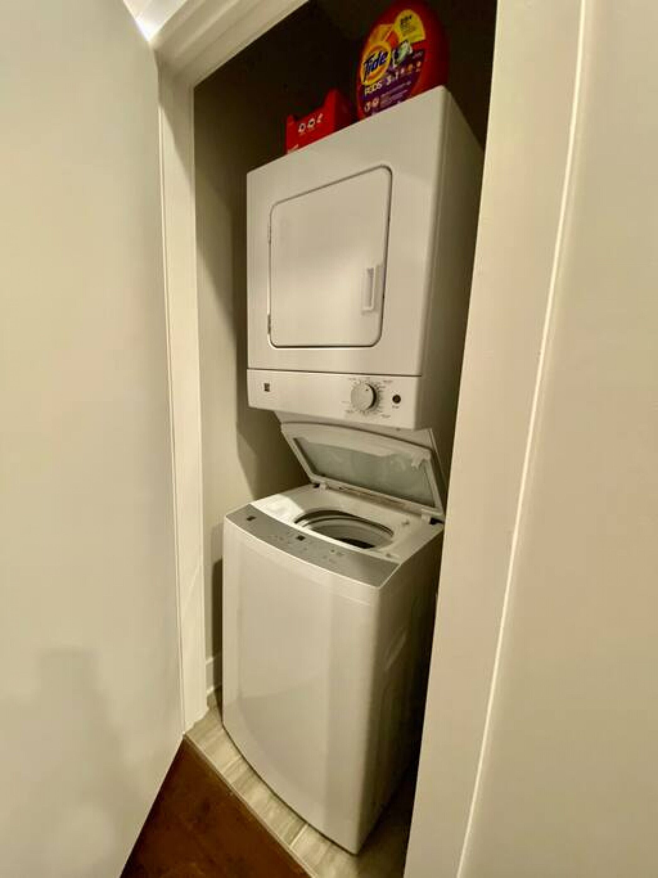 A compact, white front-loading washer and dryer unit in a narrow hallway with a brown floor.