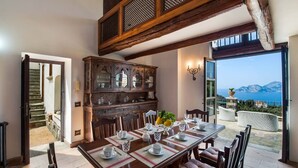 Zona pranzo con tavolo in legno apparecchiato per sei persone, circondato da poltrone imbottite. Sullo sfondo, una grande credenza in legno con vetrate.