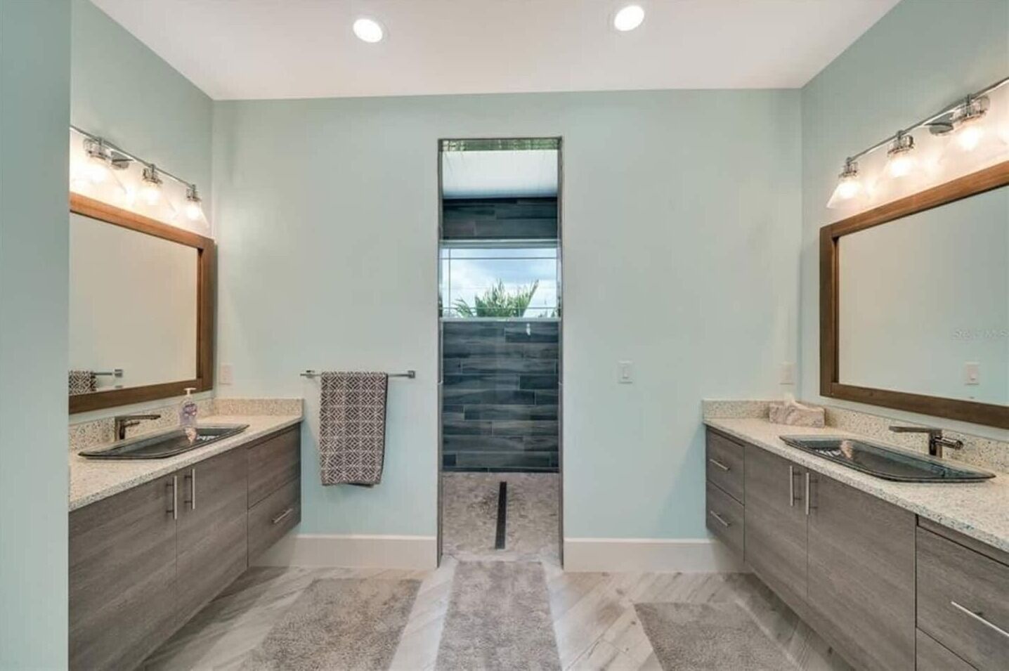 A modern bathroom with two sinks, a large mirror, and a view through a glass door to an outdoor area.