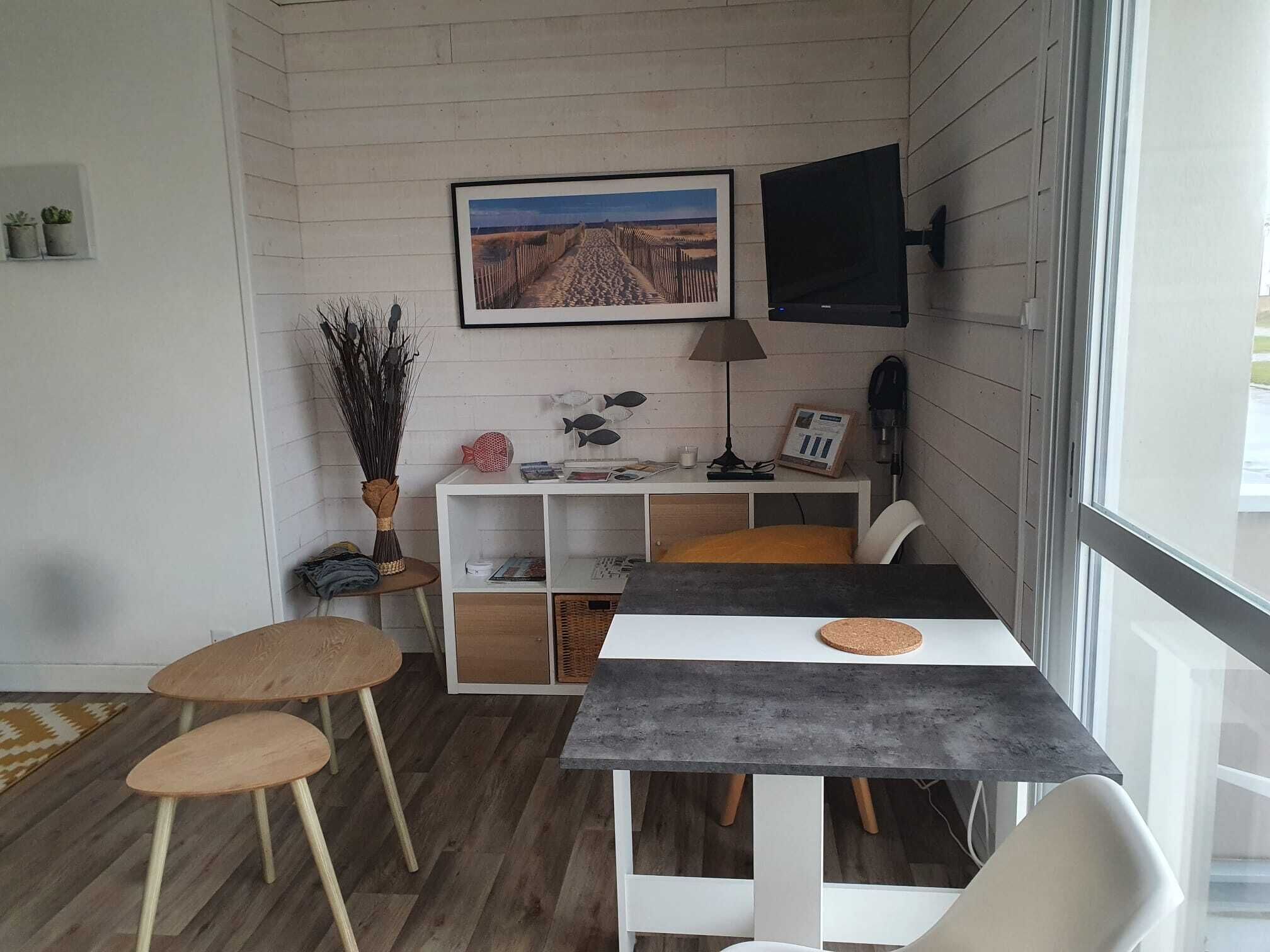 Une petite pièce agréable, dotée d’un plancher en bois, d’un bureau blanc avec un dessous de verre en liège, d’une chaise blanche et d’un téléviseur fixé au mur.