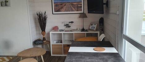 Une petite pièce agréable, dotée d’un plancher en bois, d’un bureau blanc avec un dessous de verre en liège, d’une chaise blanche et d’un téléviseur fixé au mur.