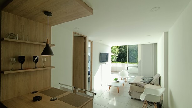 A modern living space with a wooden dining area, a white sofa, and a large window providing a view of greenery outside. - Apartamento (Medellín)