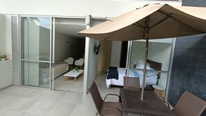 A modern outdoor patio with a table and chairs, a large umbrella, and a sliding glass door leading to a bedroom with a bed and a nightstand. - Apartamento (Medellín)