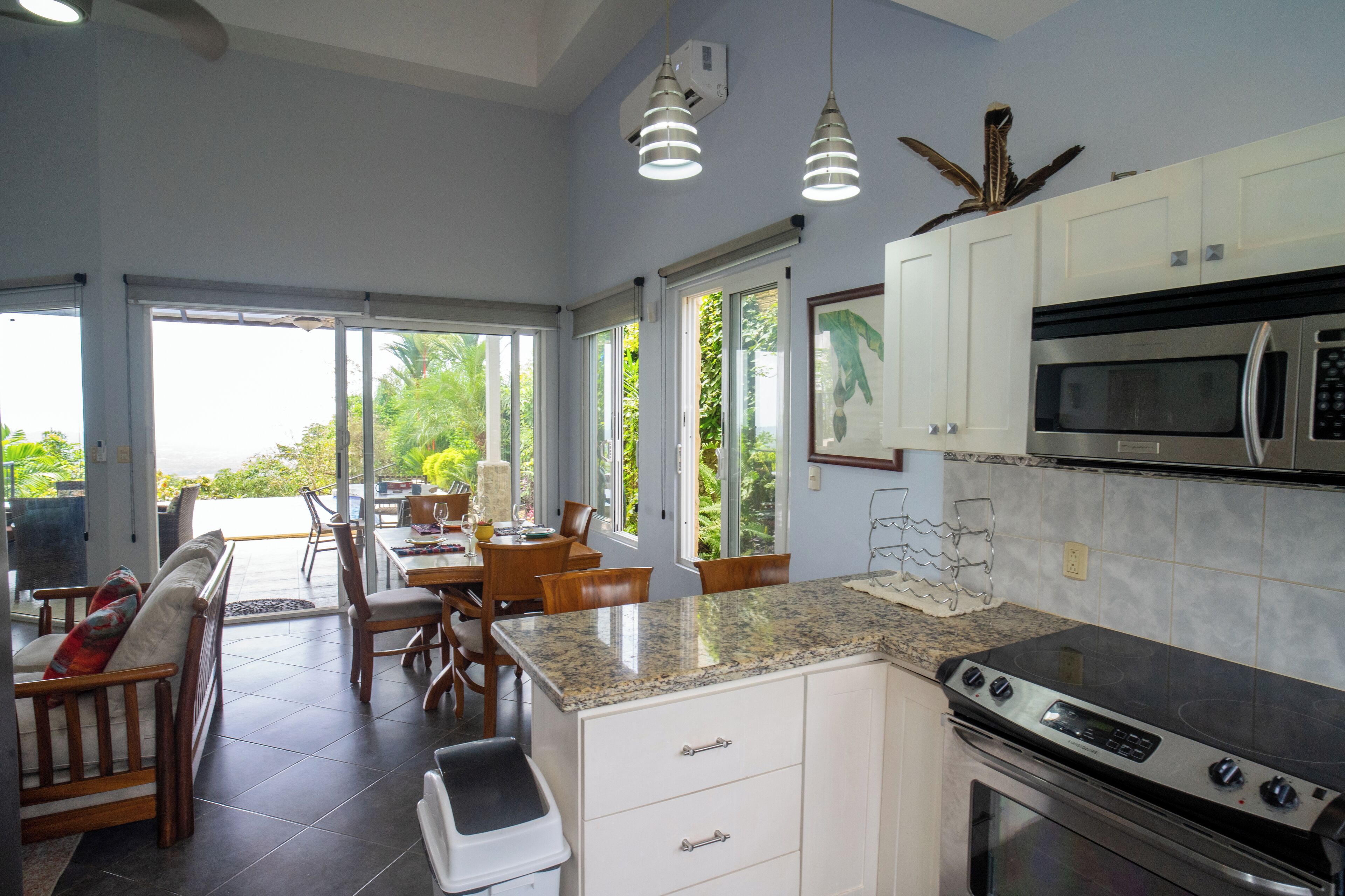 A modern kitchen with white cabinets, a granite countertop, and stainless steel appliances. There is a dining area with wooden furniture and large windows offering a view of greenery.