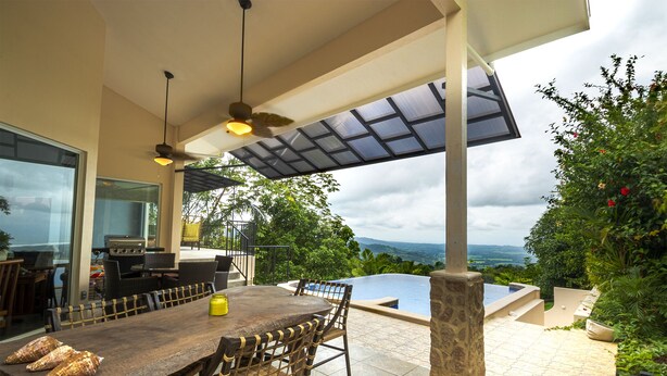 Un espace extérieur aménagé pour les repas, avec une table et des chaises en bois, un barbecue et une vue sur une piscine et un paysage verdoyant.