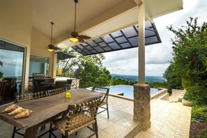Un espace extérieur aménagé pour les repas, avec une table et des chaises en bois, un barbecue et une vue sur une piscine et un paysage verdoyant.