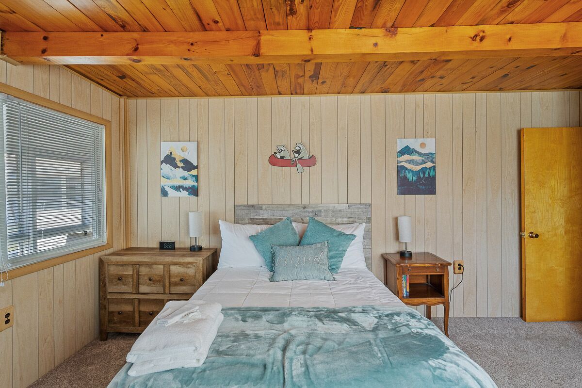 Une chambre à coucher aux murs lambrés, comprenant un lit, deux tables de chevet, une fenêtre avec des stores et une porte.