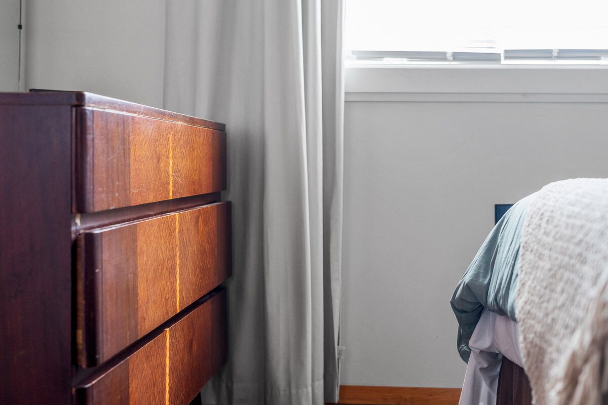 Une commode en bois avec des tiroirs, une fenêtre avec des rideaux et un lit avec une couverture.