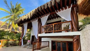 A two-story building with a thatched roof, wooden balcony railings, and a stone-paved patio. - Casa Pirata (Isla Holbox)