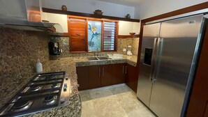 A kitchen with granite countertops, a stainless steel refrigerator, a gas stove, wooden cabinets, and a window with blinds. - Casa Pirata (Isla Holbox)