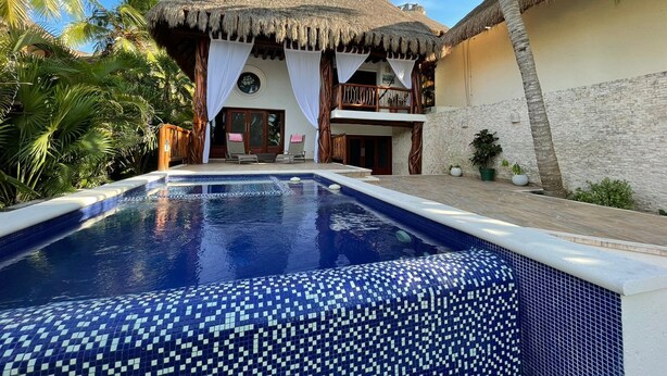 A pool with a mosaic tile pattern, a thatched-roof structure, and a balcony with white curtains. - Casa Pirata (Isla Holbox)