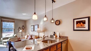 A kitchen with granite countertops, wooden cabinets, a stainless steel sink, pendant lights, a clock, framed pictures, and a window with blinds. - River Run by Wyndham Vacation Rentals (Keystone)