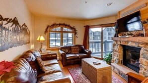 A living room with a stone fireplace, leather sofas, a television, and a large window with a view of buildings. - River Run by Wyndham Vacation Rentals (Keystone)