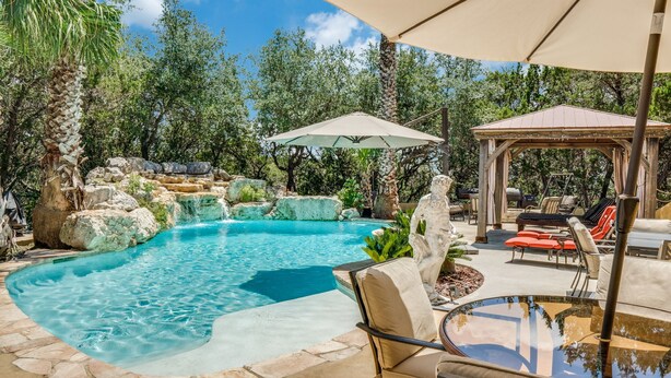 A swimming pool with a waterfall feature, surrounded by stone steps and greenery. There are lounge chairs and a gazebo in the background.