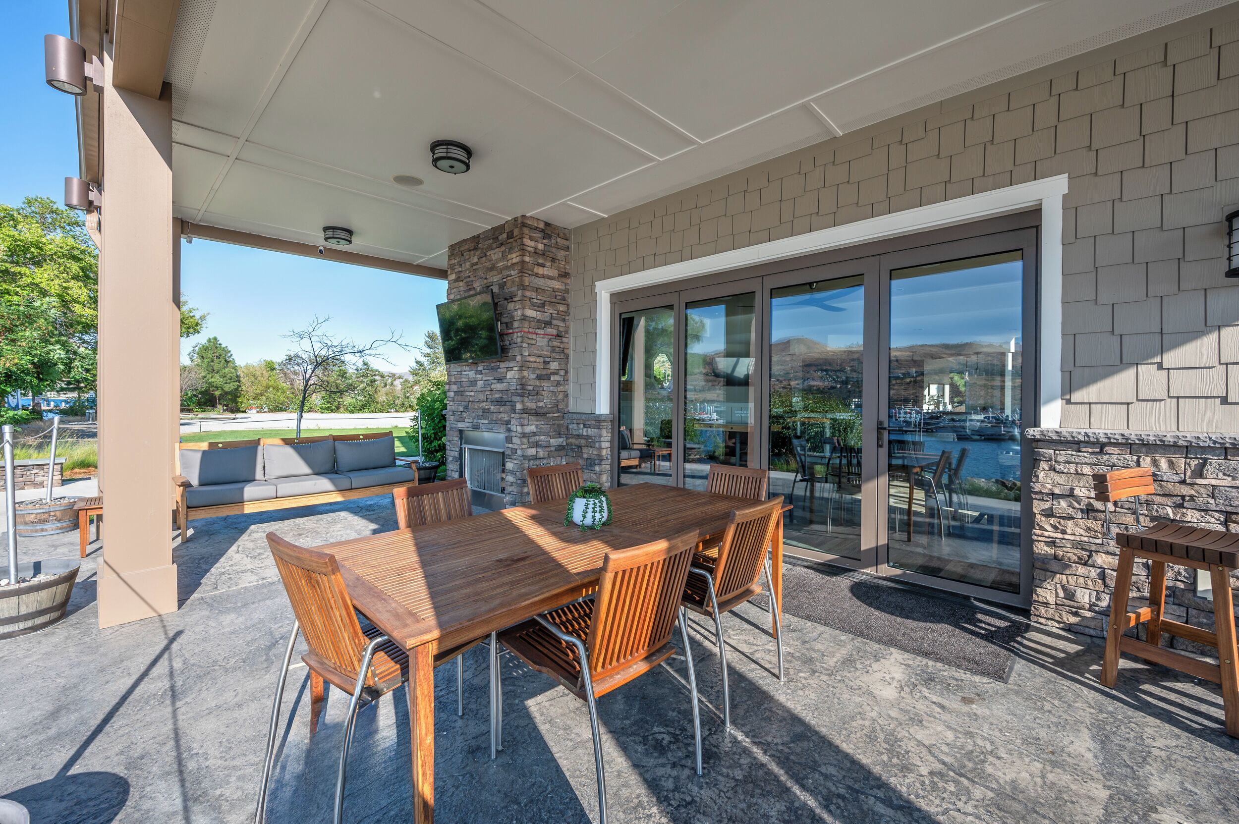 An outdoor patio with a wooden table and chairs, a stone fireplace, and a glass door leading to a view of a marina.