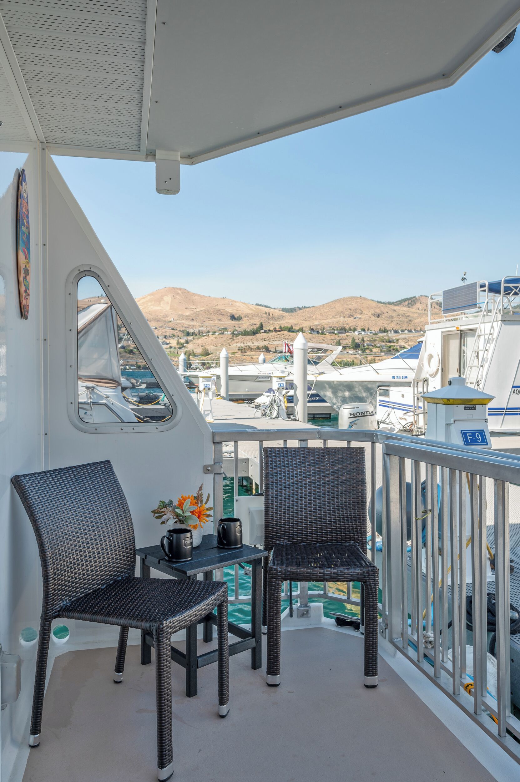 A balcony with wicker chairs and a table, overlooking a marina with boats and hills in the background.