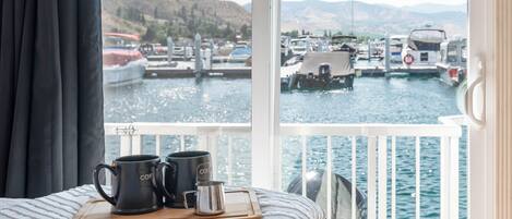 Una cama con vista a un puerto deportivo y barcos, una bandeja de madera con tazas, y una barandilla blanca.
