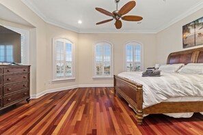 Un dormitorio con piso de madera, una cama con cabecera, un ventilador de techo y tres ventanas con contraventanas.
