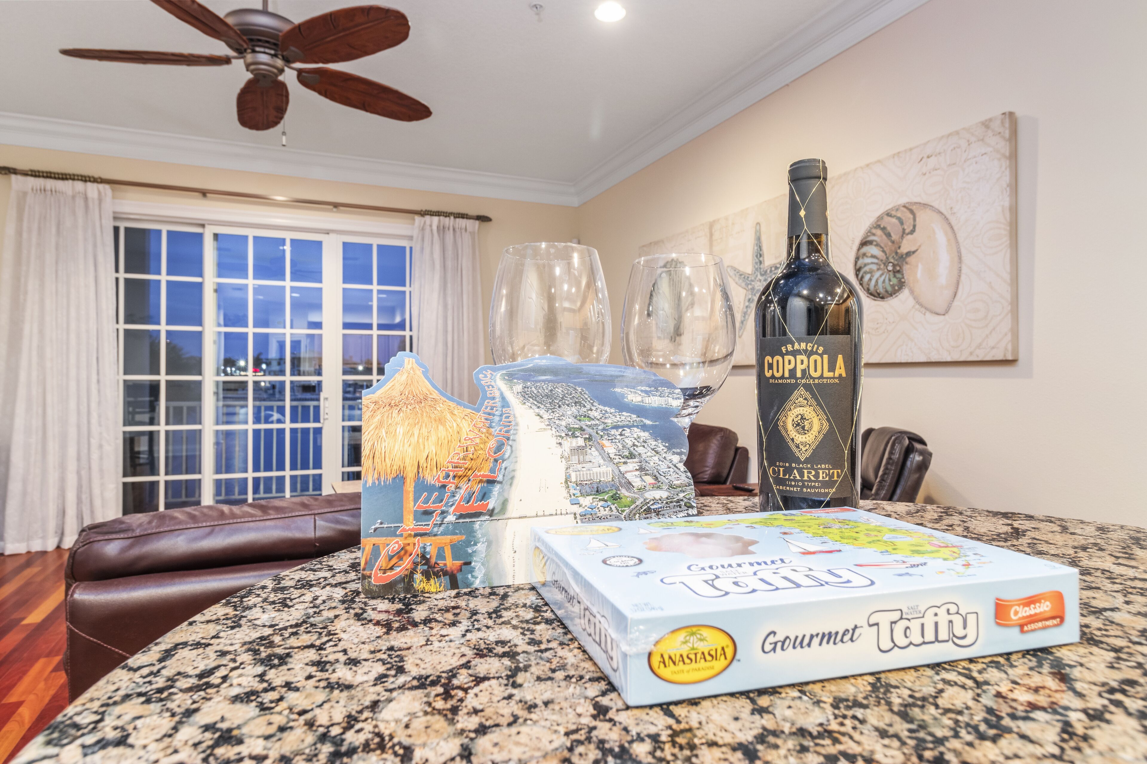A dining area with a table set for two, featuring a bottle of Francis Coppola's Claret, a box of Gourmet Taffy, and a decorative item resembling a beach scene.