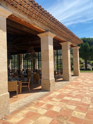 Un patio al aire libre con columnas de piedra, mesas y sillas de madera, bajo un techo de tejas.