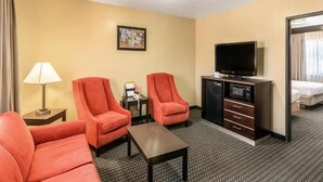 Habitación de hotel con dos sillones rojos, una mesita, un televisor en un mueble bajo, un cuadro en la pared y una cama al fondo.