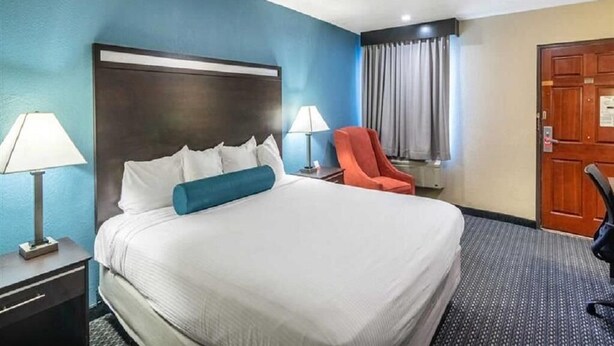 Habitación de hotel con una cama grande, dos lámparas de noche, una silla roja, una puerta de madera y una ventana con cortinas.