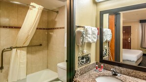 Baño de hotel con ducha, inodoro, lavabo y un espejo grande que refleja un dormitorio.