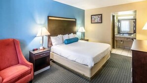 Habitación de hotel con cama, un sillón rojo, una mesita de noche y un baño visible a través de una puerta abierta.