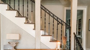 A staircase with a wrought iron railing, a wooden console table with a vase and candle, and a view of a room with a cabinet and a mirror. - Spacious Historic Midtown Memphis Chateau Minutes from Everything  (Memphis)