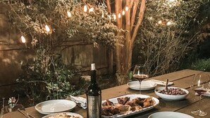 An outdoor dining table set with plates, bowls, and a bottle of wine, surrounded by greenery and string lights. - Spacious Historic Midtown Memphis Chateau Minutes from Everything  (Memphis)