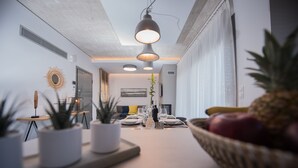 A modern dining area with a long table set for two, potted plants, and a fruit basket. - Emmeleia mare new building maisonette with pool, seaview, 3 min from the beach  (Αγία Πελαγία)