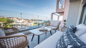 A balcony with wicker furniture, a glass table, and a view of the sea and buildings. - Emmeleia mare new building maisonette with pool, seaview, 3 min from the beach  (Αγία Πελαγία)