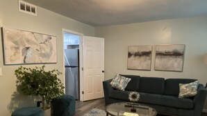 A living room with a dark green sofa, two abstract paintings, a glass coffee table, a potted plant, and a wooden floor. - Newly Renovated Cozy/Modern Pet-Friendly Villa for the entire family to enjoy. (Hilton Head Island)