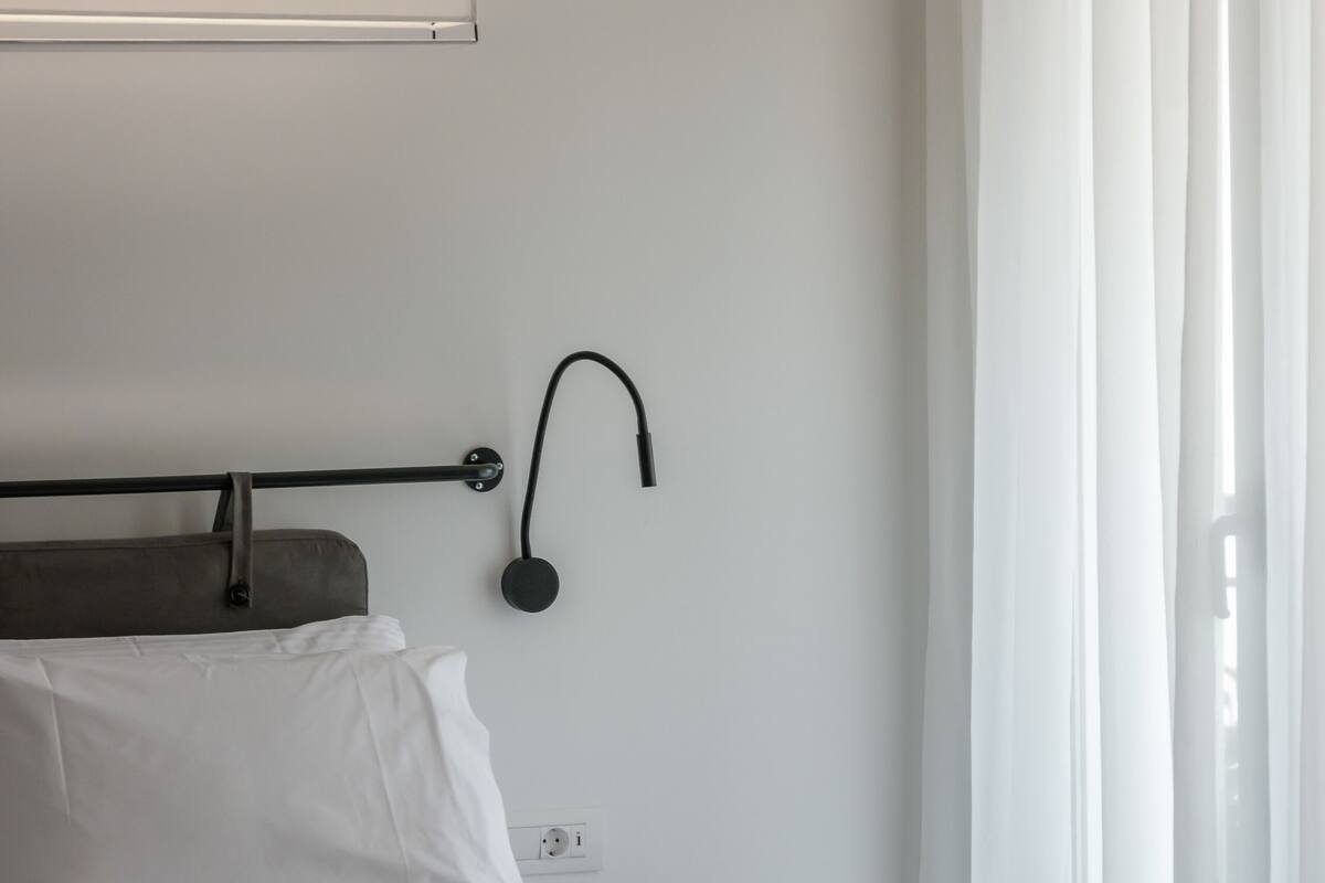 Une chambre d’hôtel moderne, équipée d’un lit, d’une table de chevet, d’une lampe fixée au mur et d’une fenêtre avec des rideaux légers.