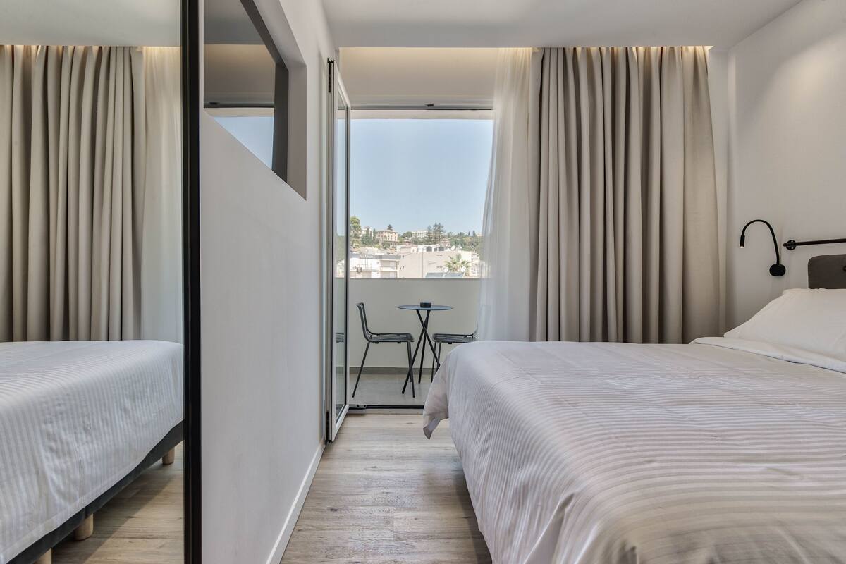 Une chambre d’hôtel avec deux lits, un balcon avec une table et des chaises, et une vue sur la ville.