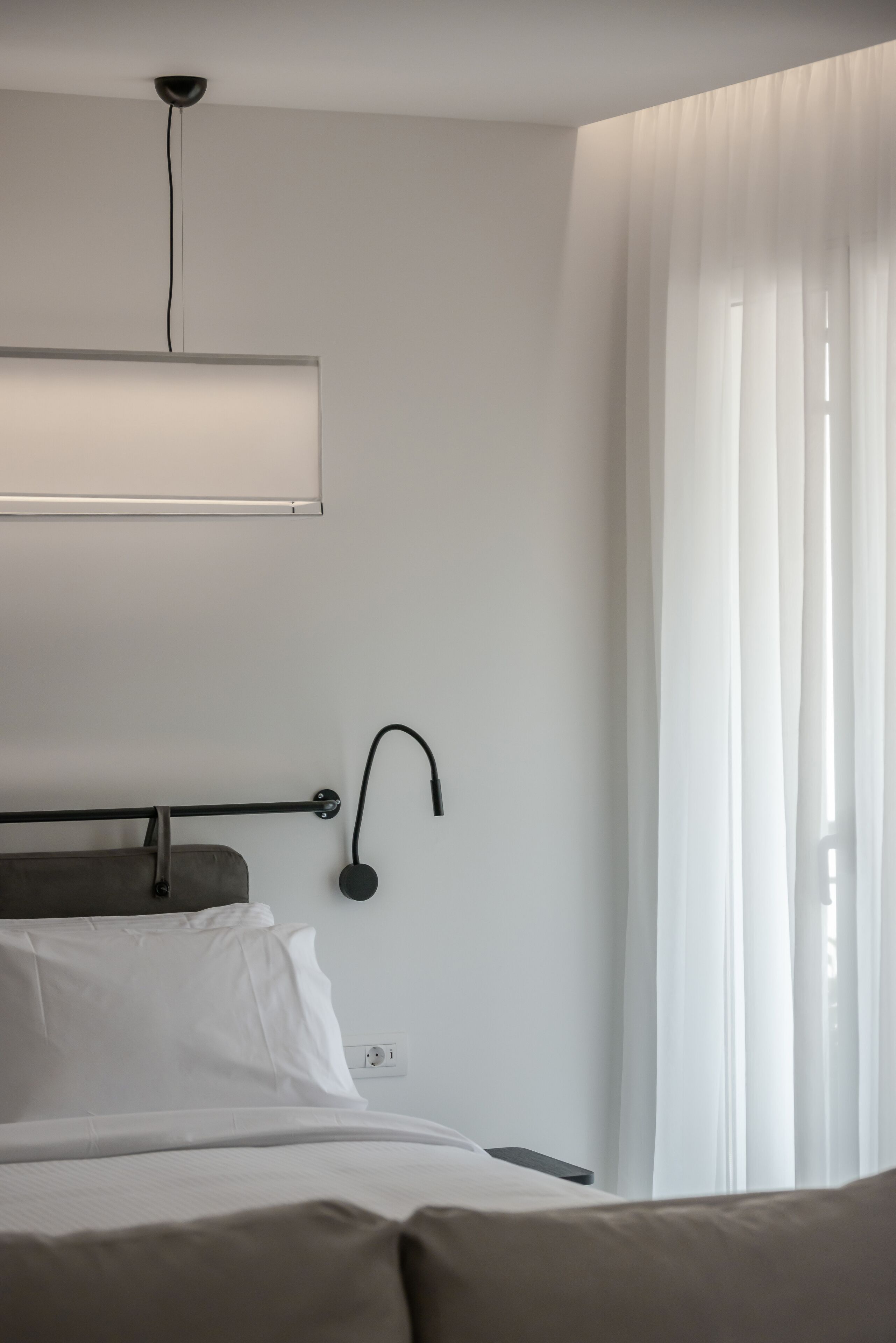 A hotel room with a bed, a bedside table, a wall-mounted lamp, and a window with sheer curtains.