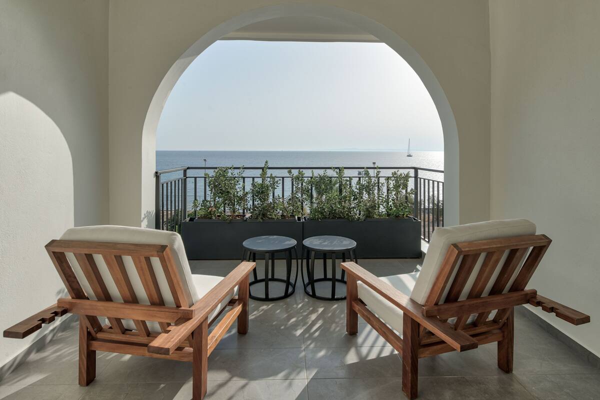 Un balcon avec deux chaises en bois et deux petites tables rondes, offrant une vue sur la mer, agrémenté d’une balustrade et de verdure.