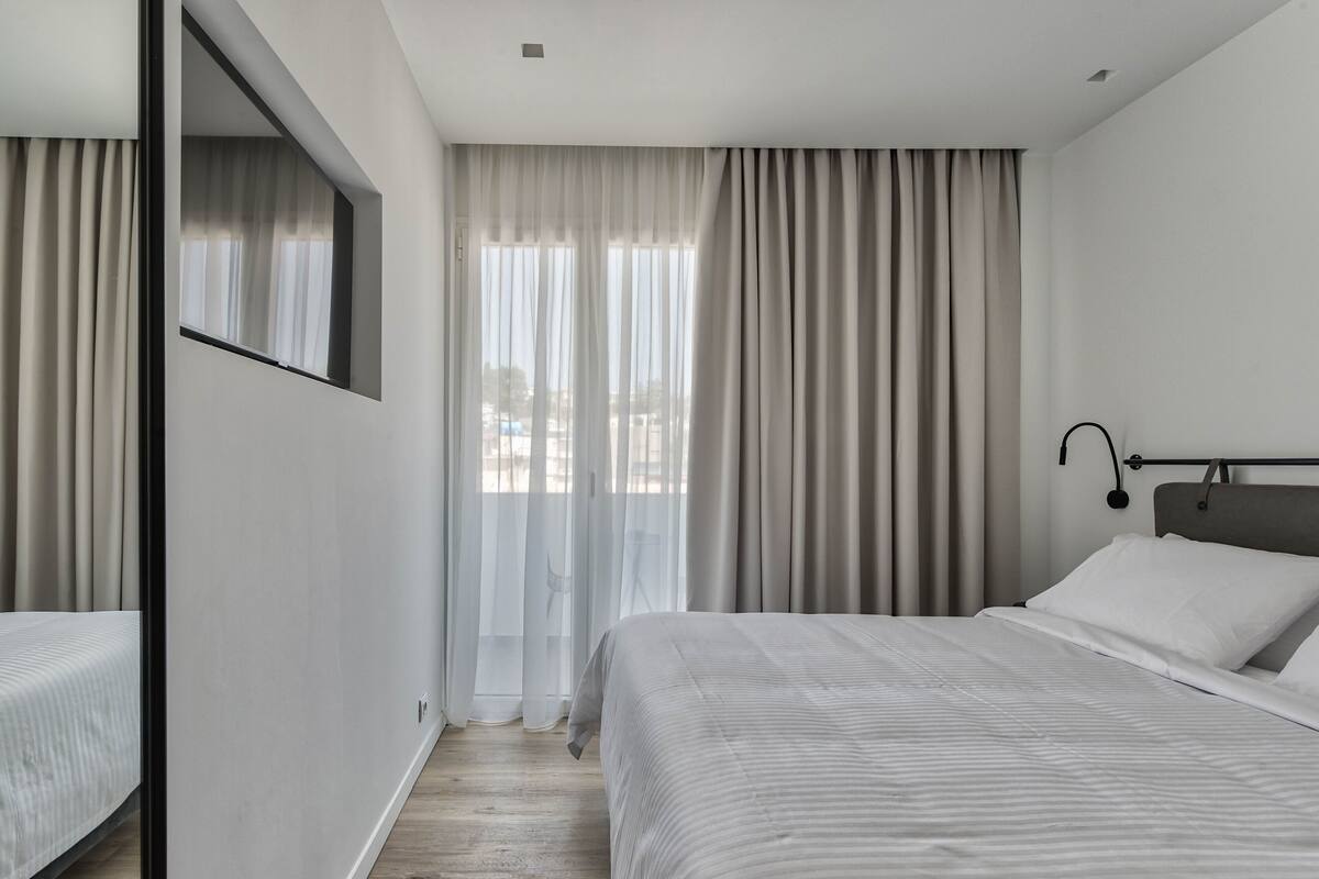 Une chambre d’hôtel moderne, dotée d’un grand lit, d’une table de chevet, d’un miroir et d’une fenêtre avec des rideaux légers.