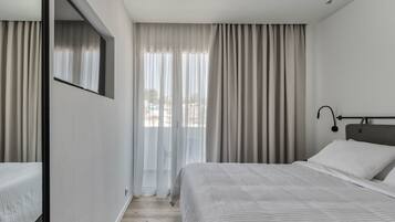 Ein modernes Hotelzimmer mit einem großen Bett, einem Nachttisch, einem Spiegel und einem Fenster mit transparenten Vorhängen.