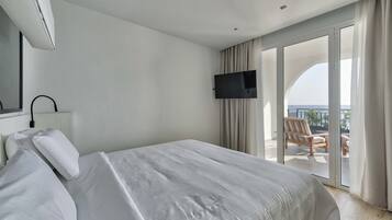 A hotel room with a large bed, a flat-screen TV mounted on the wall, and a sliding glass door leading to a balcony with a view of the sky and a chair.