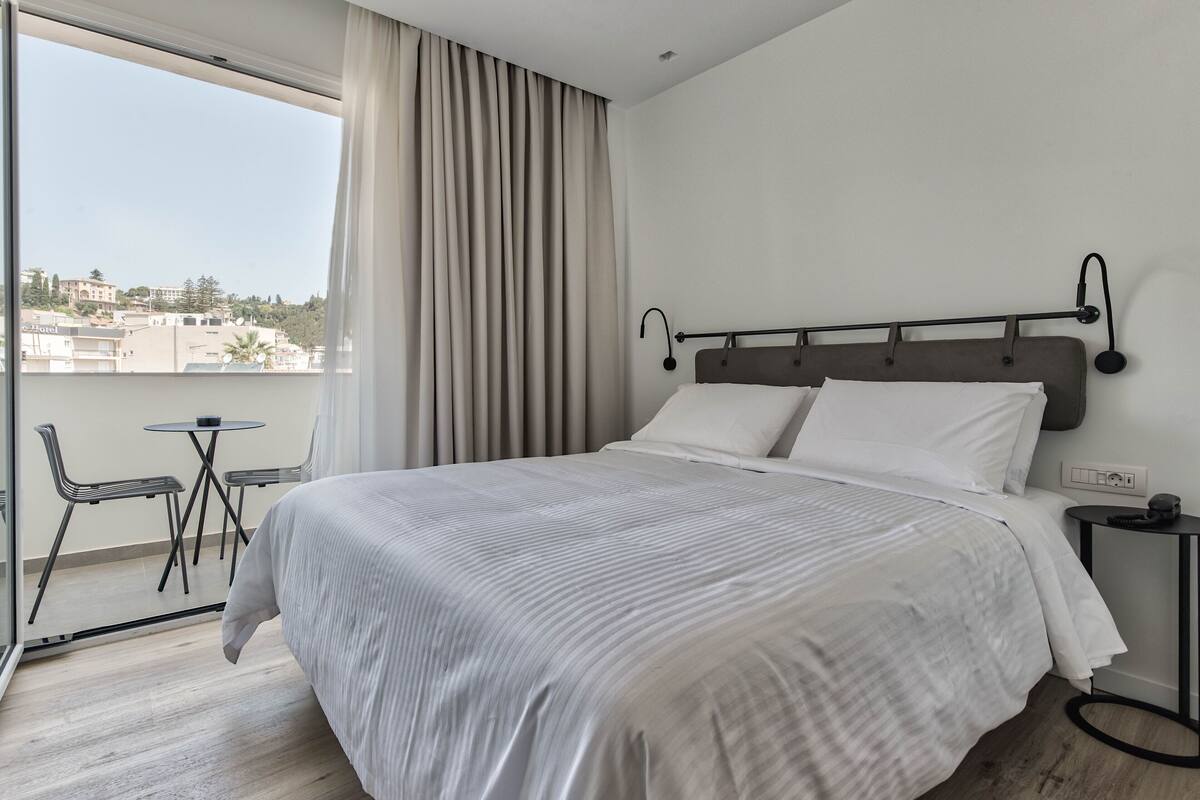 Une chambre d’hôtel avec un lit, une petite table, deux chaises, une fenêtre avec des rideaux et une vue sur des immeubles.