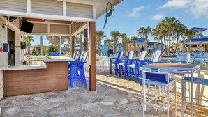 A covered outdoor area with a bar, blue and white striped chairs, and a poolside setting with palm trees and buildings in the background.