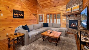 L’intérieur d’une cabane en rondins, avec une cheminée en pierre, un plafond en bois et un grand canapé d’angle.