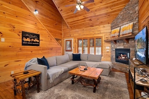 A log cabin interior with a stone fireplace, wooden ceiling, and a large sectional sofa. - In the Woods - log cabin w/hot tub, game room, BBQ (Gatlinburg)