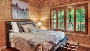 Une chambre à coucher dans un chalet en bois, avec un lit, des oreillers, un tableau, une table de chevet, une lampe et une fenêtre avec des stores.