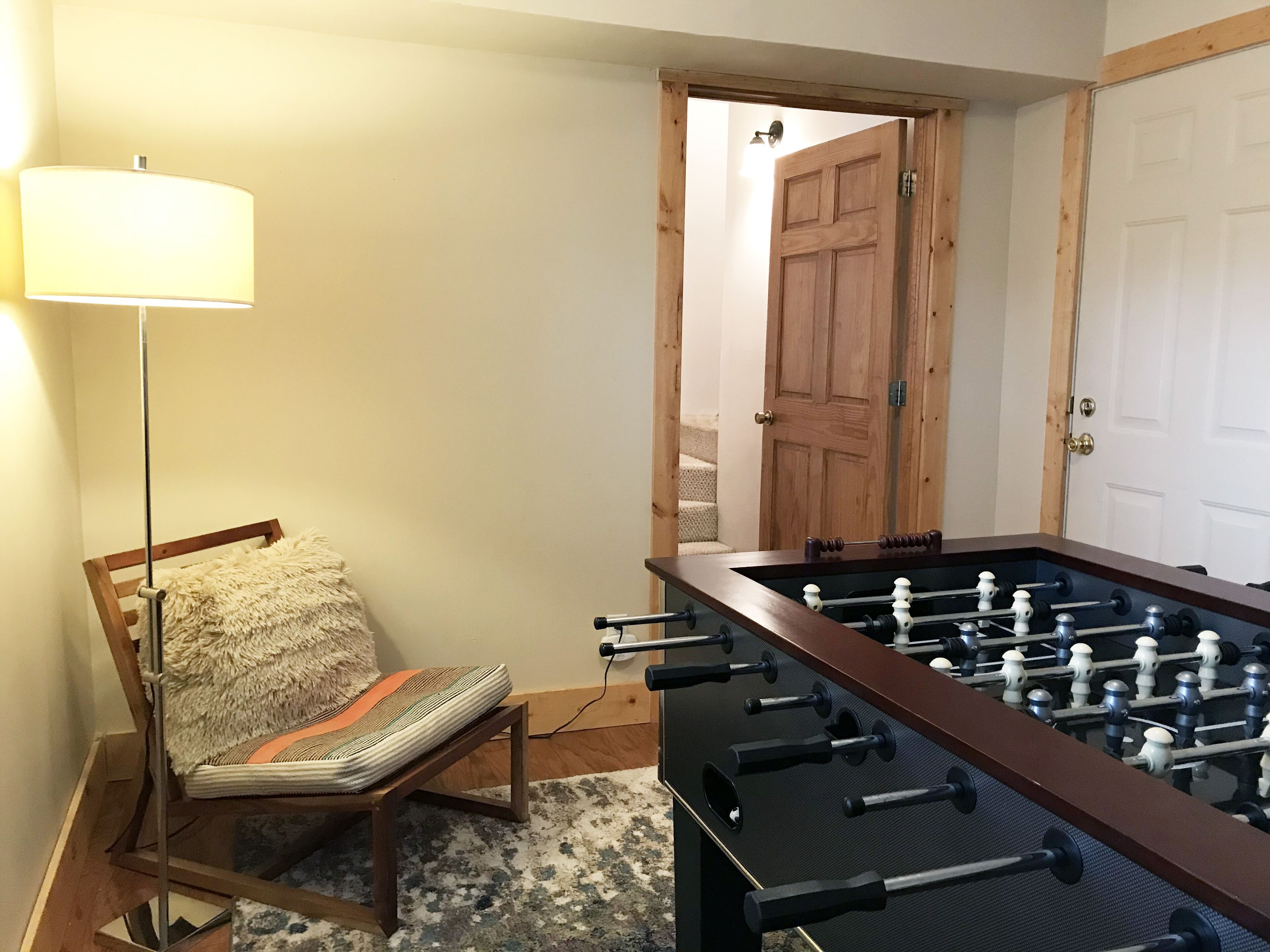 Een kamer met een tafelvoetbaltafel, een lamp, een stoel met kussen en een deur.