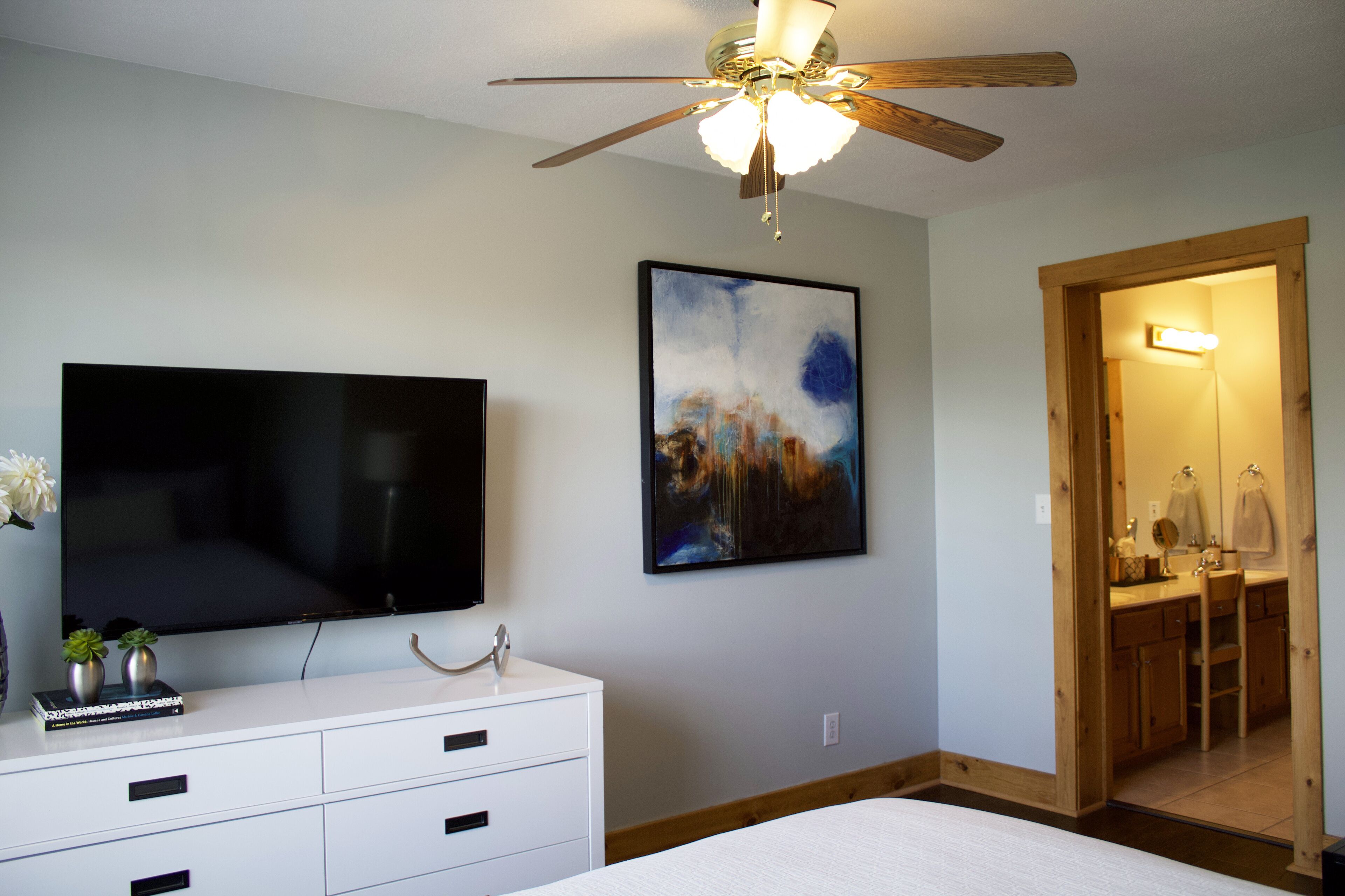 A bedroom with a bed, a TV, a ceiling fan, a painting, and a bathroom visible through an open door.