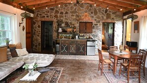 A rustic interior with a stone wall, wooden ceiling beams, a kitchen area, a dining table, and a seating area with a patterned rug. - Scottland Yard Farm's - Upscale Rustic Garden House (Brooktondale)